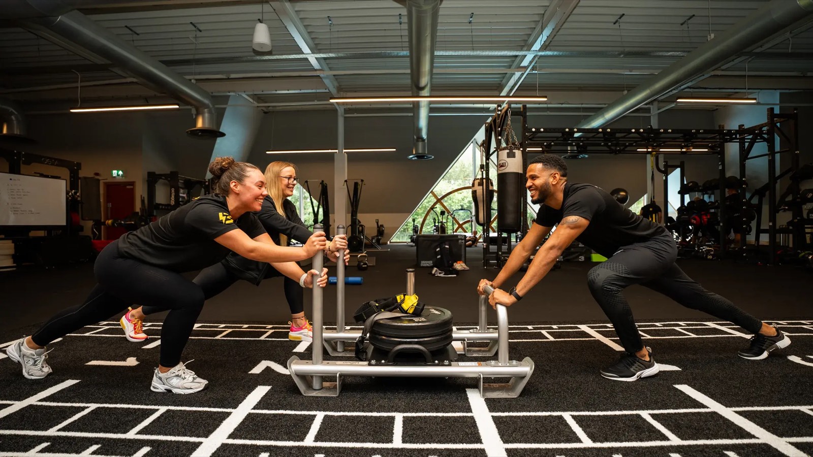A coach and two smiling gym members pushing a weight sled together during a workout